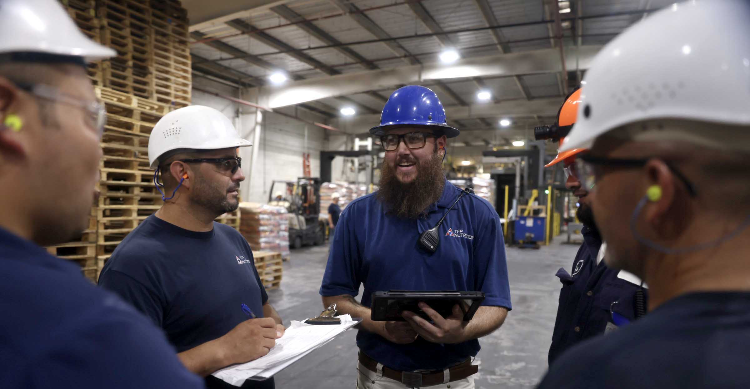 A group of warehouse workers in discussion with papers and a tablet.