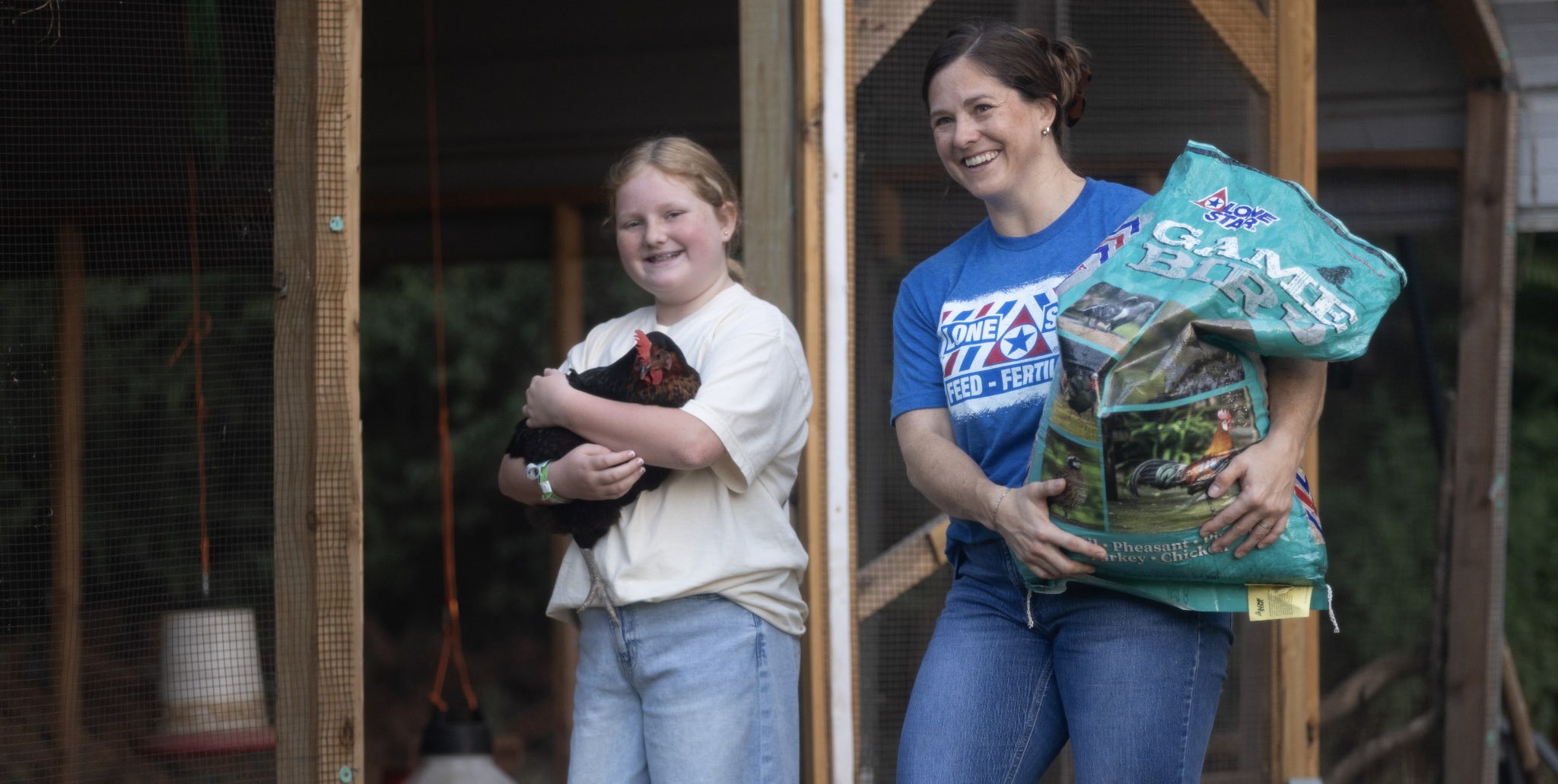 woman carrying bag of feed alongside a girl carrying a chicken
